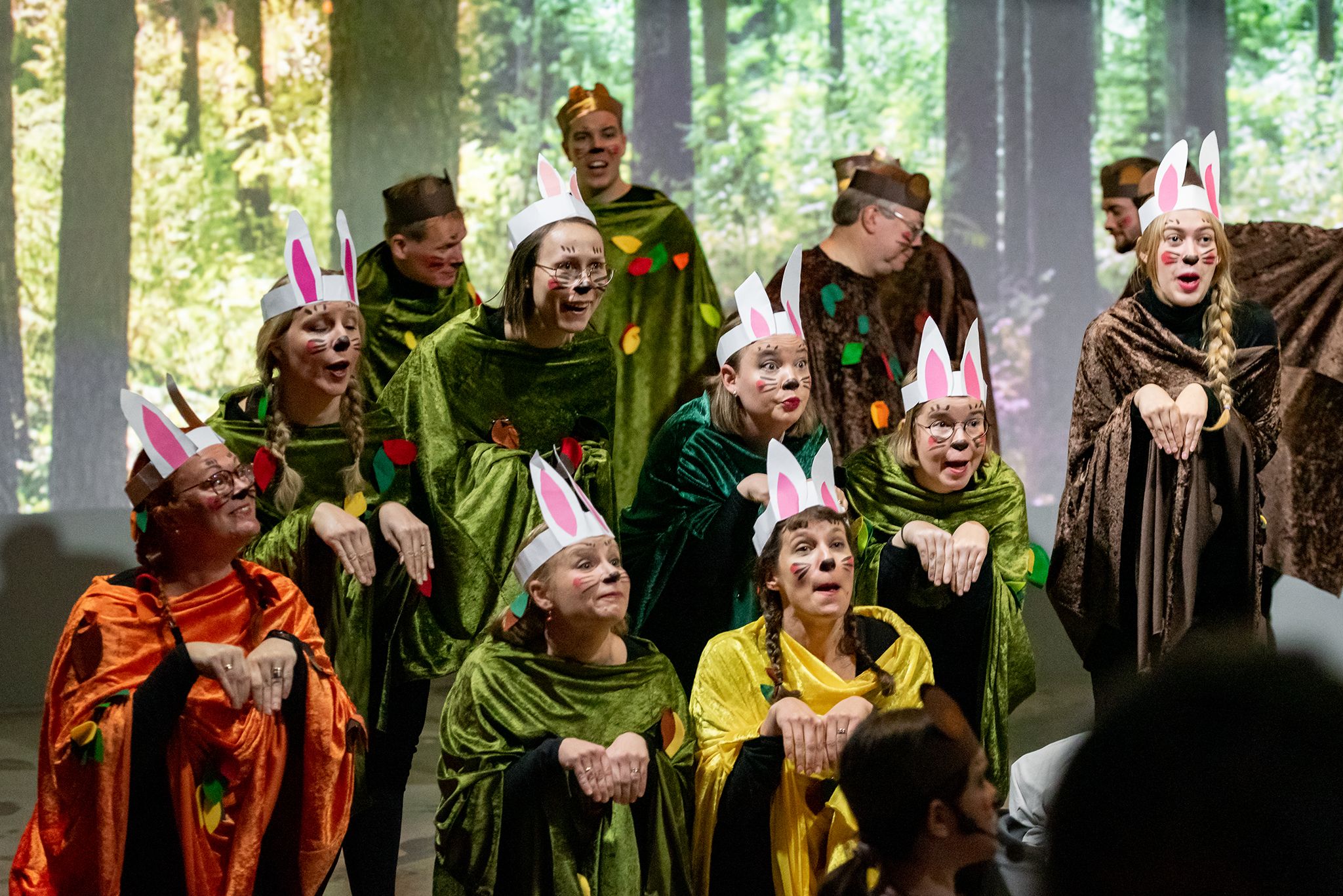 Kuorolaisia vihreisiin ja ruskeisiin kaapuihin pukeutuneena ja pahviset pupukorvat päässä, poseeraavat jäniksinä lavalla 