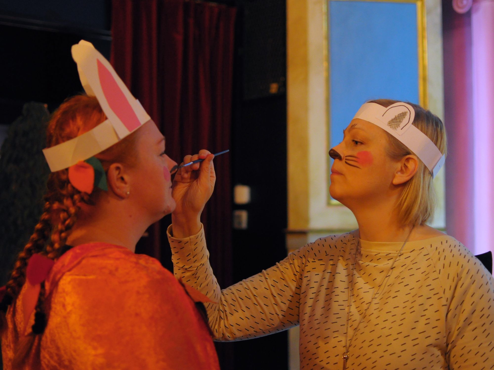 Kuoronjohtaja maalaa kuorolaiselle kasvomaalia, molemmat ovat pukeutuneet lastenkonserttia varten