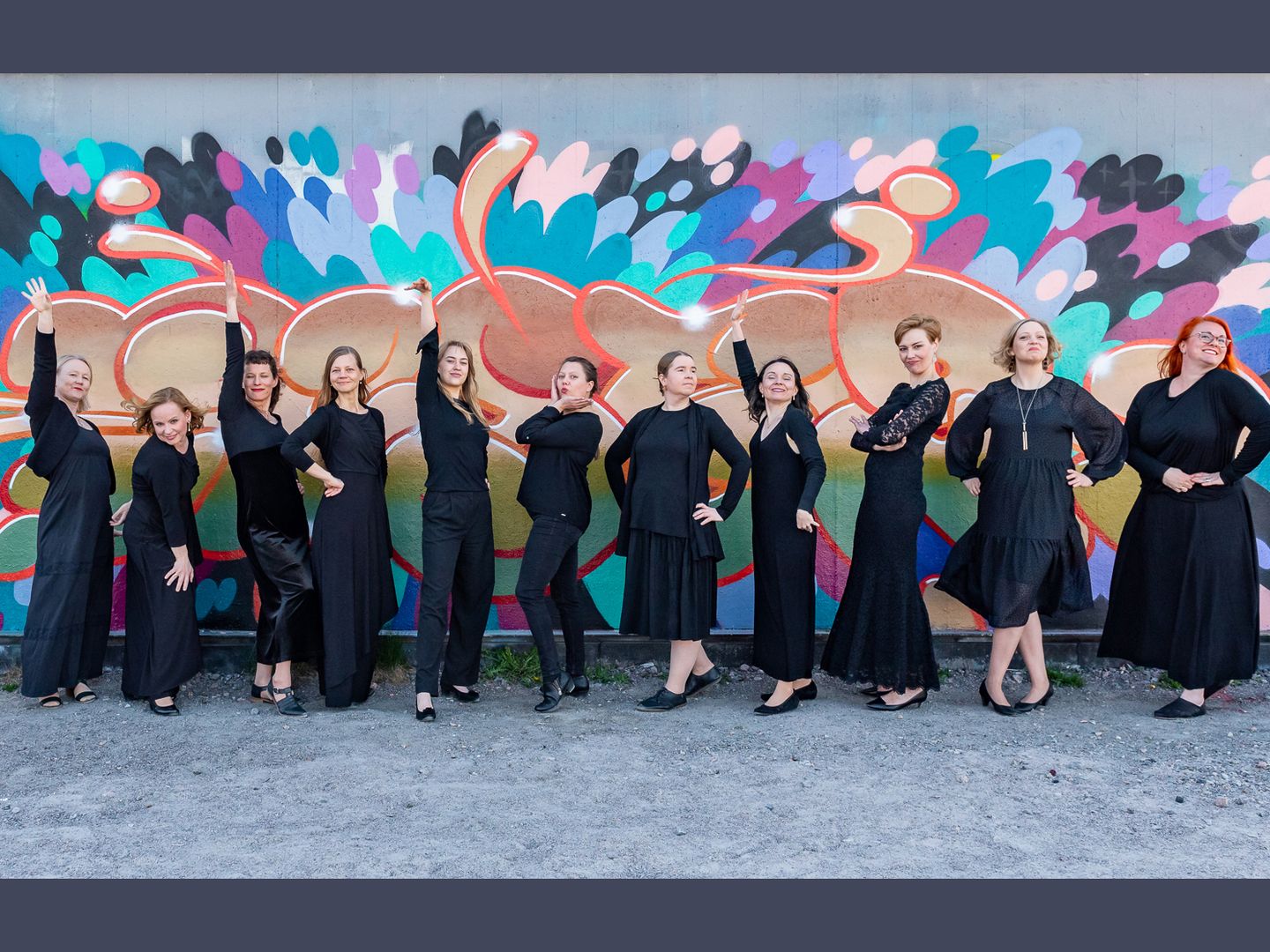 Mustiinpukeutuneet kuoron naislaulajat poseeraavat dramaattisin, mutta koomisin elein graffitiseinän edessä