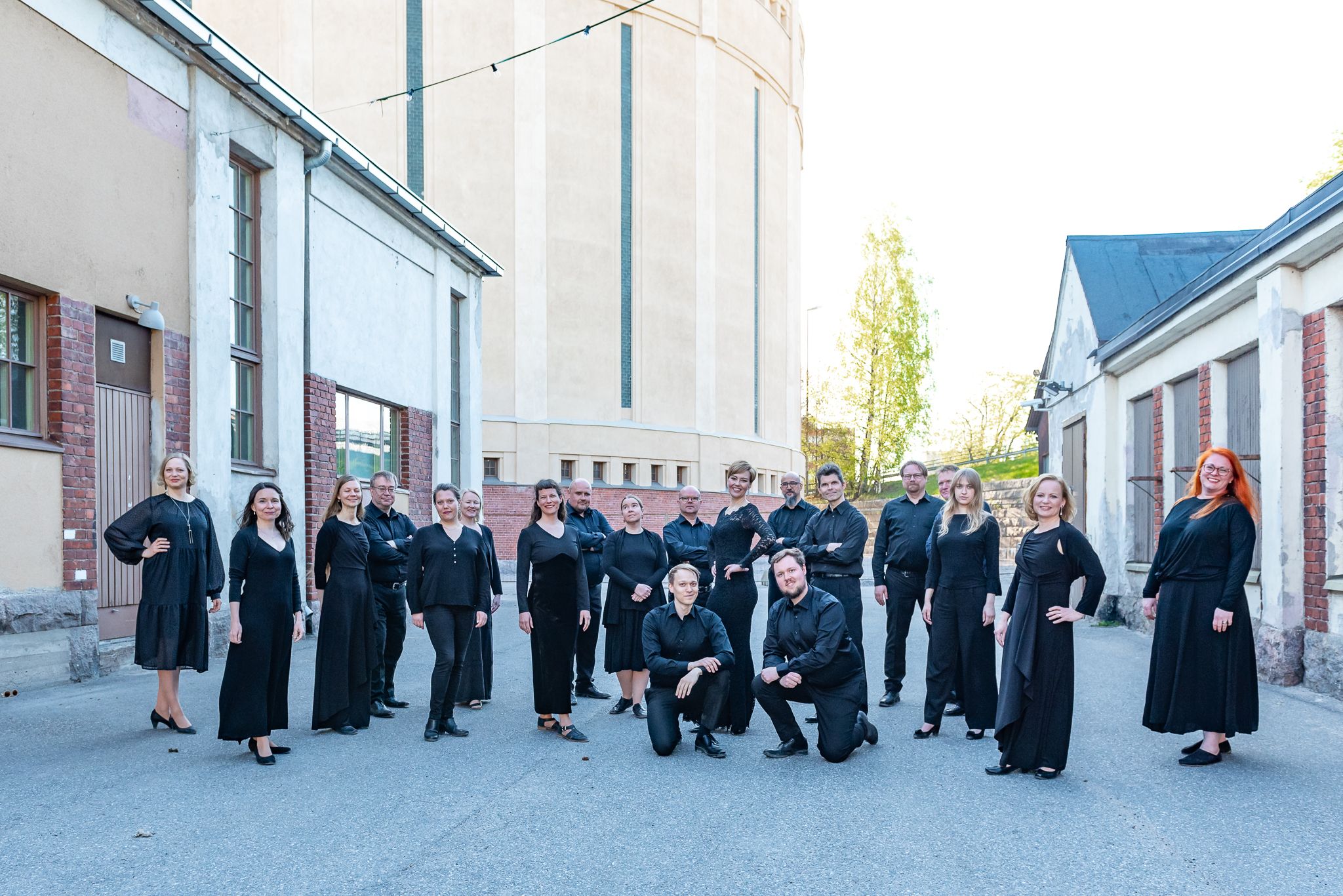 Mustiinpukeutuneet kuorolaiset seisovat väljässä ryhmäkuvassa vanhojen tehdasrakennusten välissä