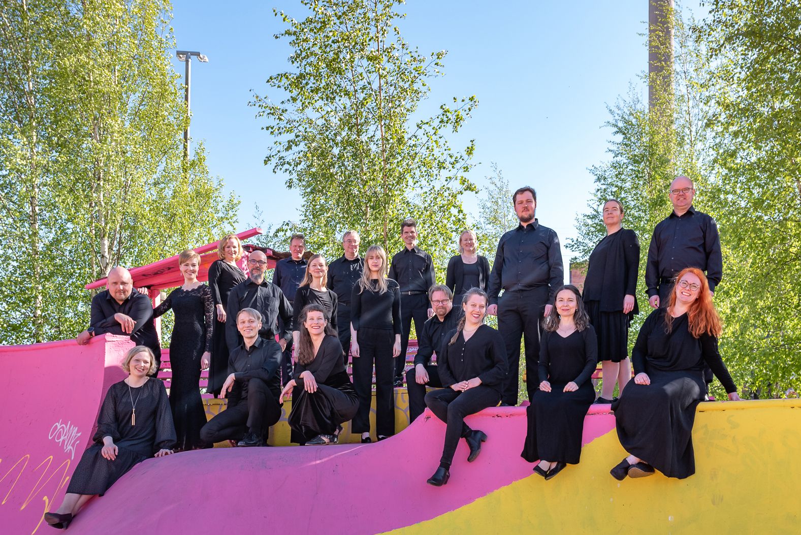 Mustiin pukeutuneet kuorolaiset rennossa ryhmäkuvassa skeittipuiston laidalla aurinkoisena päivänä